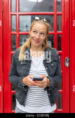 Femme souriante tenant le smartphone en regardant loin tout en se tenant contre cabine téléphonique Banque D'Images