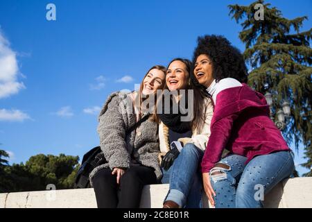 Des amies gaies qui regardent loin tout en étant assis sur le mur de soutènement contre le ciel bleu dans le parc Banque D'Images