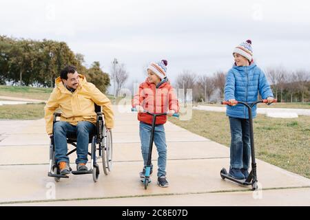 Père assis en fauteuil roulant pendant que les garçons se déplacent en scooter sentier dans le parc Banque D'Images