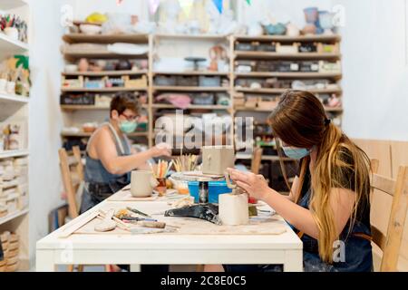 Femmes portant des masques de fabrication de céramique sur banc dans l'atelier Banque D'Images