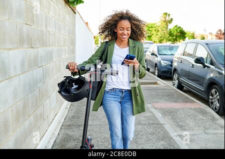 Une jeune femme heureuse utilisant un smartphone tout en marchant avec de l'électricité pousser le scooter sur le trottoir Banque D'Images