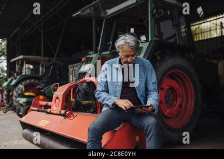 Homme âgé utilisant une tablette sur une ferme avec un tracteur en marche grange Banque D'Images