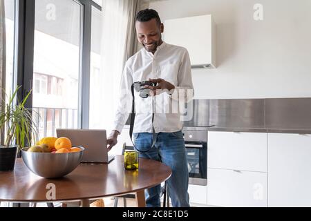 Homme souriant utilisant un ordinateur portable et un appareil photo à la maison Banque D'Images