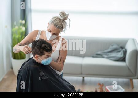 Coiffeur femme portant un masque coupant les cheveux de garçon à la maison pendant couvre-feu Banque D'Images