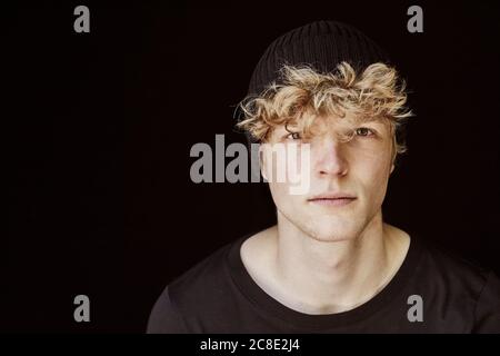 Portrait d'un jeune homme aux cheveux blonds bouclés portant du noir capuchon sur fond noir Banque D'Images