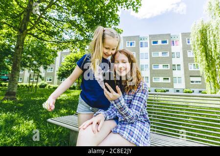 Des sœurs souriantes partageant un smartphone sur un banc dans le parc Banque D'Images