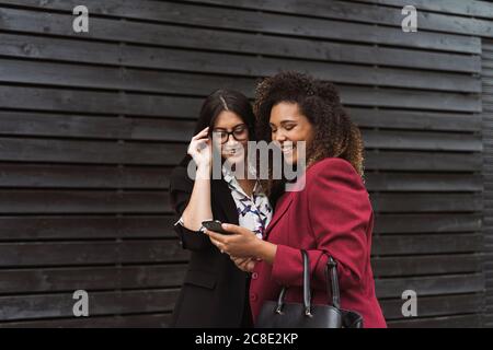 Une femme entrepreneure heureuse qui partage son smartphone tout en se tenant contre le noir mur Banque D'Images