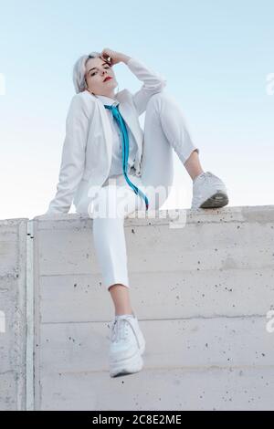 Jeune femme fraîche portant un costume blanc assis sur un mur de soutènement contre le ciel dégagé Banque D'Images