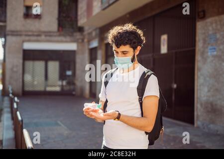 Jeune homme portant un masque se lavant les mains avec de l'assainisseur en étant debout contre le bâtiment Banque D'Images