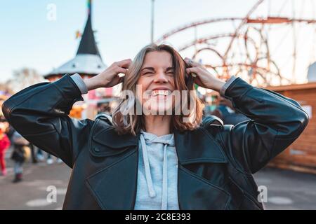 Gros plan d'une jeune femme gaie avec les mains dans les cheveux debout au parc d'attractions au coucher du soleil Banque D'Images