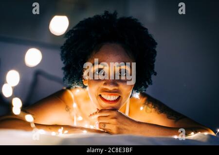 Gros plan d'une femme souriante avec des guirlandes lumineuses illuminées qui se détendent lit à la maison Banque D'Images