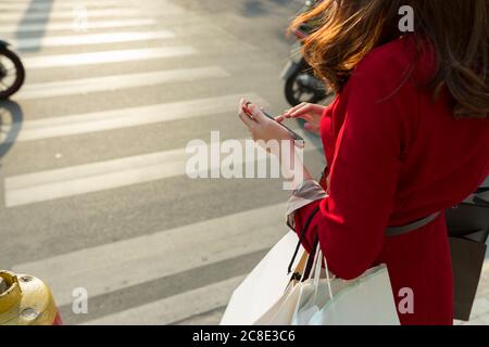 Jeune femme portant des sacs d'achats tenant un smartphone lors de son passage route en ville Banque D'Images