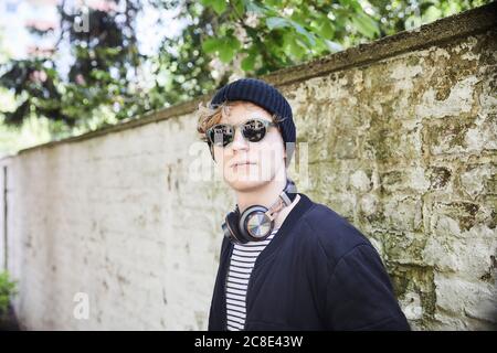 Portrait d'un jeune homme avec casque portant un capuchon et des lunettes de soleil devant le mur abîmé Banque D'Images