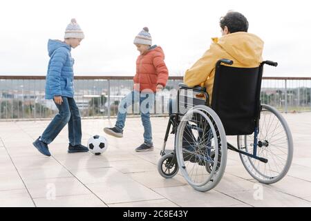 Homme assis en fauteuil roulant regardant des fils jouer avec le ballon en stationnement dans un ciel dégagé Banque D'Images