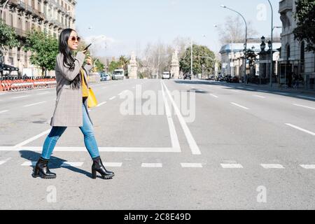 Jeune femme élégante parlant sur smartphone en traversant la route en ville Banque D'Images