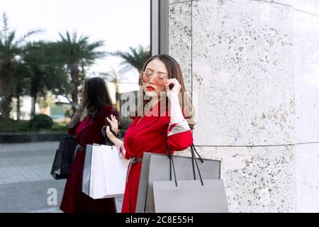 Jeune femme portant des lunettes de soleil portant des sacs à provisions tout en étant debout bâtiment moderne Banque D'Images