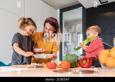 Une mère et une fille souriantes préparent la nourriture pendant que bébé fille est assis sur l'île de cuisine Banque D'Images