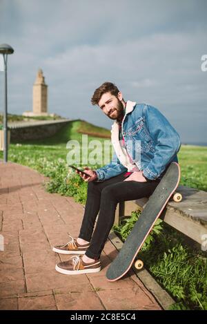 Jeune homme charmant utilisant un smartphone assis avec un skateboard sur le siège Banque D'Images