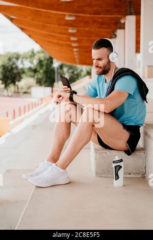 Athlète masculin avec casque, smartphone et montre intelligente ayant une pause assis sur la tribune dans le stade Banque D'Images