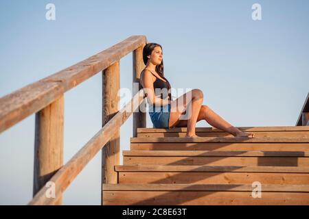 Adolescente bien réfléchie assise sur des marches en bois bleu clair ciel Banque D'Images