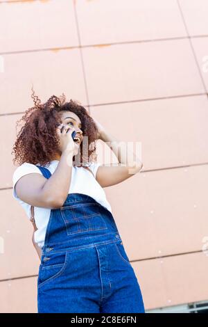 Jeune femme gaie avec des cheveux afro parlant au téléphone intelligent en se tenant contre le mur Banque D'Images