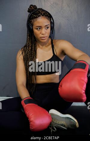 Athlète féminine fatiguée portant des gants rouges écoutant de la musique pendant qu'elle est assise contre le mur dans la salle de gym Banque D'Images