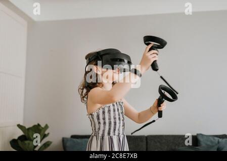 Fille portant le simulateur de réalité virtuelle jouant avec des joysticks à la maison Banque D'Images