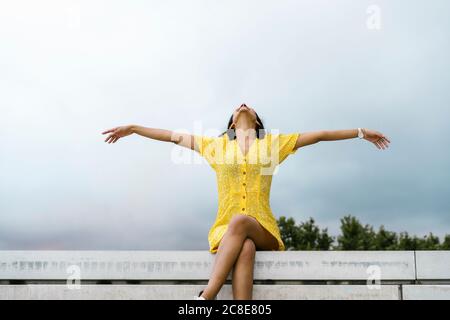 Jeune femme insouciante assise avec les bras étirés sur le mur de retenue contre le ciel Banque D'Images