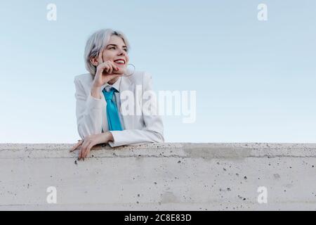 Jeune femme souriante portant un costume blanc debout près d'un mur de soutènement contre le ciel dégagé Banque D'Images