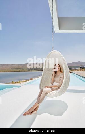 Bonne femme assise dans une chaise suspendue au-dessus de la piscine parlant sur le téléphone Banque D'Images