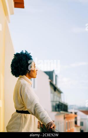 Femme attentionnée avec des cheveux bouclés regardant loin tout en restant debout balcon au ciel dégagé Banque D'Images