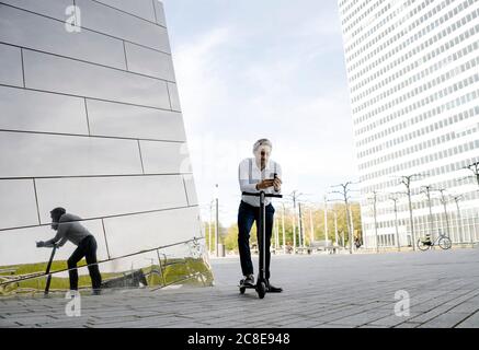 Homme d'affaires avec kick scooter et smartphone se reflète dans la façade du bâtiment Banque D'Images