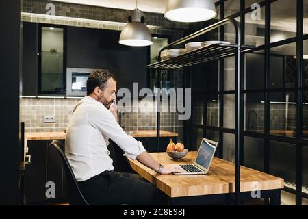 Homme d'affaires mature travaillant dans la cuisine, utilisant un ordinateur portable, parlant au téléphone Banque D'Images