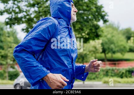 Homme âgé portant un imperméable bleu qui court contre les arbres dans le parc Banque D'Images