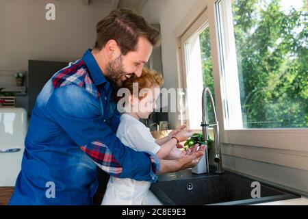 Homme souriant se lavant les mains avec son fils dans l'évier de cuisine à accueil Banque D'Images