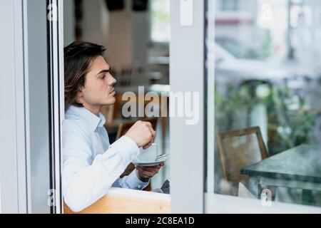 Homme d'affaires attentionné tenant une tasse de café tout en étant assis dans un café vu par la fenêtre Banque D'Images