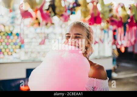 Gros plan d'une jeune femme souriante avec les yeux fermés tenant le coton bonbons au parc d'attractions Banque D'Images