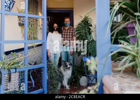 Couple portant des masques avec chien debout à l'entrée de la maison vue à travers la porte Banque D'Images
