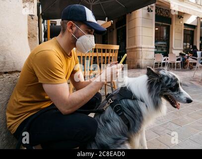Homme portant un masque de protection en attente avec son chien dans le ville à l'aide d'un smartphone Banque D'Images