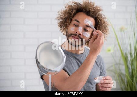 Gros plan d'un jeune homme appliquant un masque facial tout en regardant dans miroir à la maison Banque D'Images