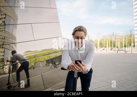 Homme d'affaires avec kick scooter et smartphone se reflète dans la façade du bâtiment Banque D'Images