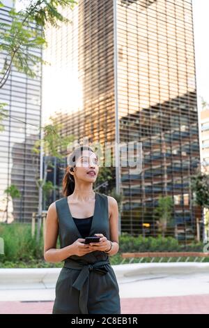 Femme d'affaires tenant un smartphone et se tenant debout contre des bâtiments modernes en ville Banque D'Images