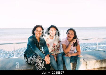 Mère souriante avec des filles assises sur le mur de soutènement contre la mer Banque D'Images