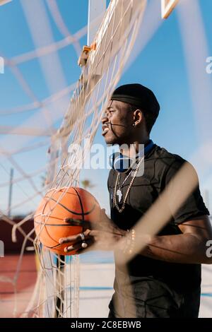 Jeune homme tenant le ballon de basket-ball en regardant loin en se tenant par le filet en cour Banque D'Images