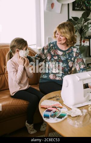 Femme souriante regardant la petite-fille portant un masque de protection maison pendant une pandémie Banque D'Images