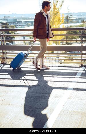 Homme d'affaires tirant ses bagages tout en marchant sur une passerelle surélevée à l'aéroport par beau temps Banque D'Images