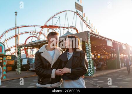 Jeune couple romantique tenant les mains tout en se tenant dans le parc d'attractions au coucher du soleil Banque D'Images