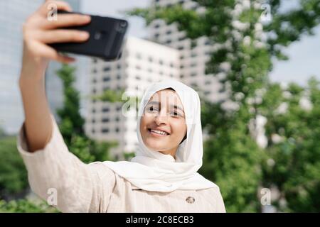 Jeune fille musulmane souriant tout en prenant le selfie à travers le smartphone en ville Banque D'Images