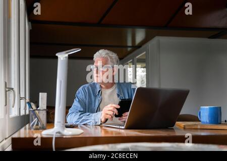 Homme senior attentionné tenant un smartphone avec un ordinateur portable sur une table à la maison Banque D'Images