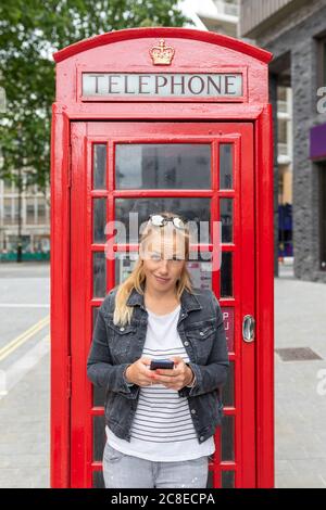 Femme adulte de taille moyenne utilisant un smartphone en se tenant contre le téléphone stand Banque D'Images